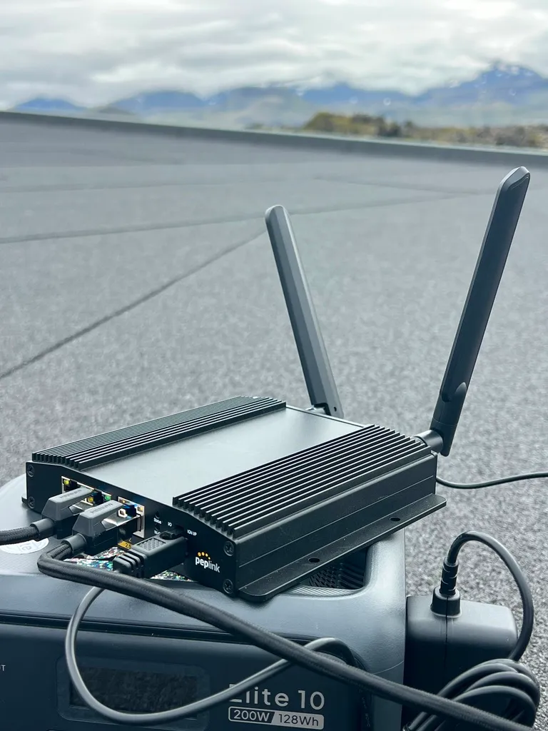 A Peplink MAX Orbit 2 router with twin antennas, its logo visible, sitting on a portable battery on a rooftop at Hellisandur, Iceland, with the mountains of the Snæfellsnes peninsula behind.