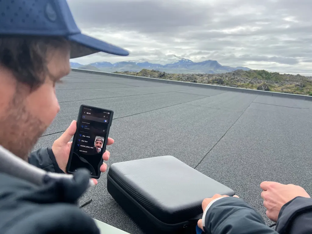 An Elite colleague on the rooftop in Iceland holding a phone showing available Wi-Fi networks, including one named Elite - Arctic, beside a weatherproof case containing a Starlink Mini.